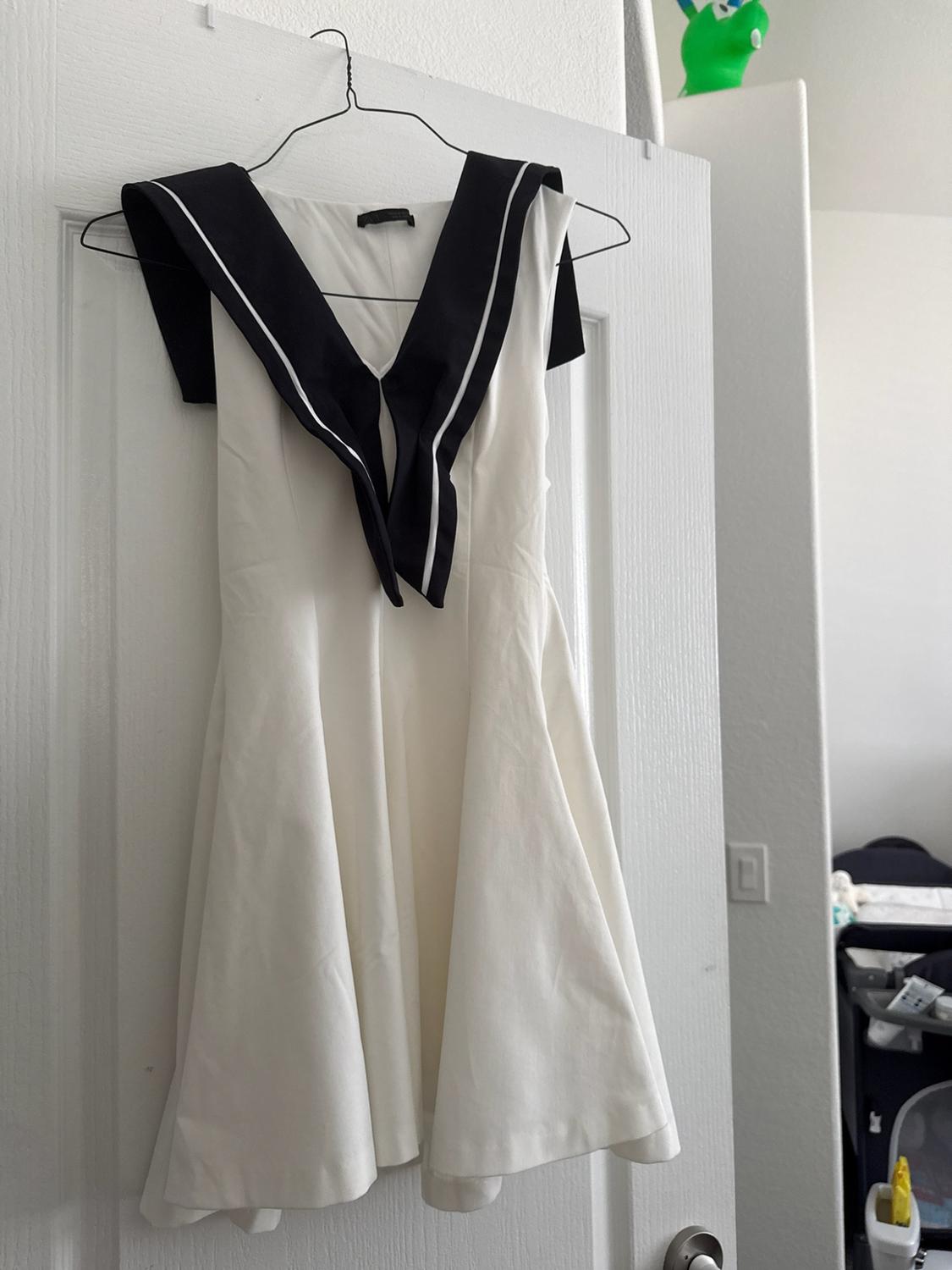 White dress with a black sailor collar featuring white stripes, hanging on a hanger on a white door.