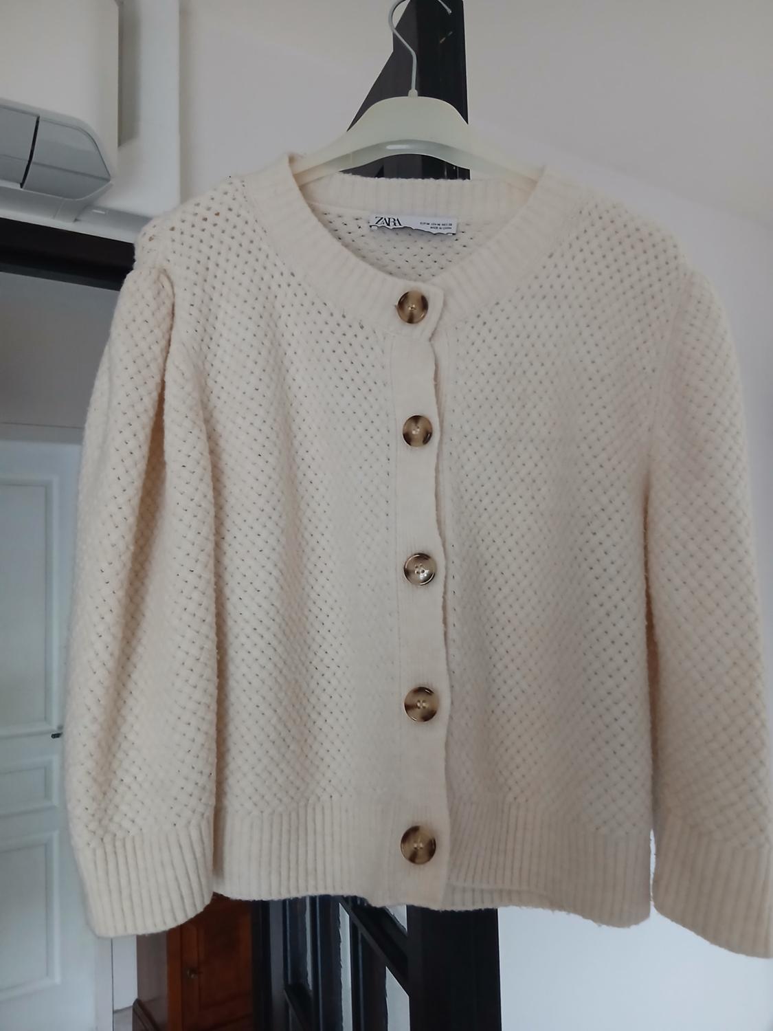 Beige knitted cardigan with large buttons hanging on a white hanger. Interior room background.