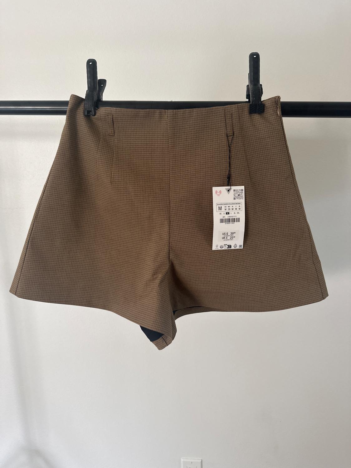 Brown shorts with a small check pattern hanging on a black hanger. Visible label with product details.
