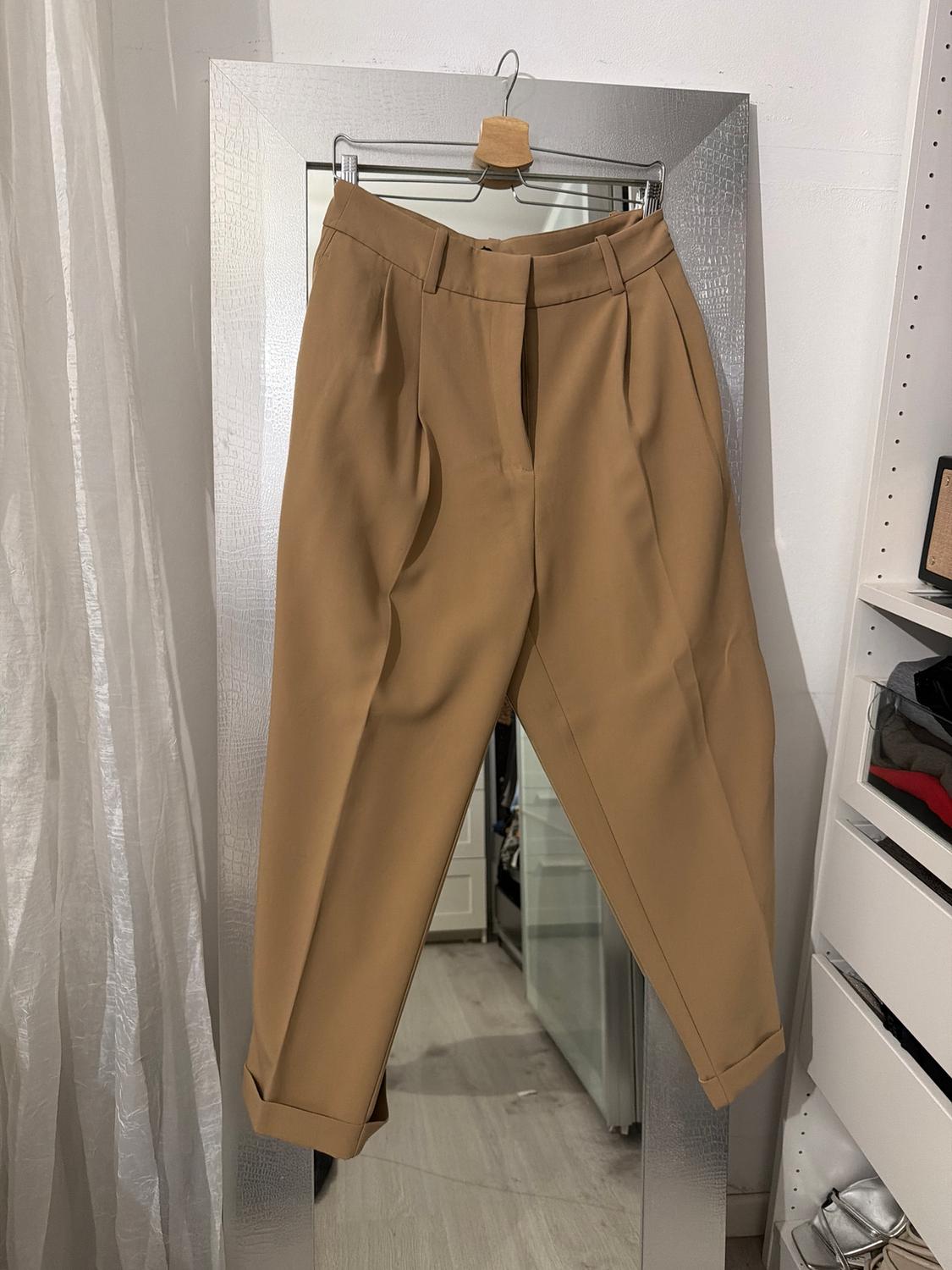 Beige trousers hanging on a hanger in front of a mirror in a wardrobe. Background with white curtain and shelves with clothes.