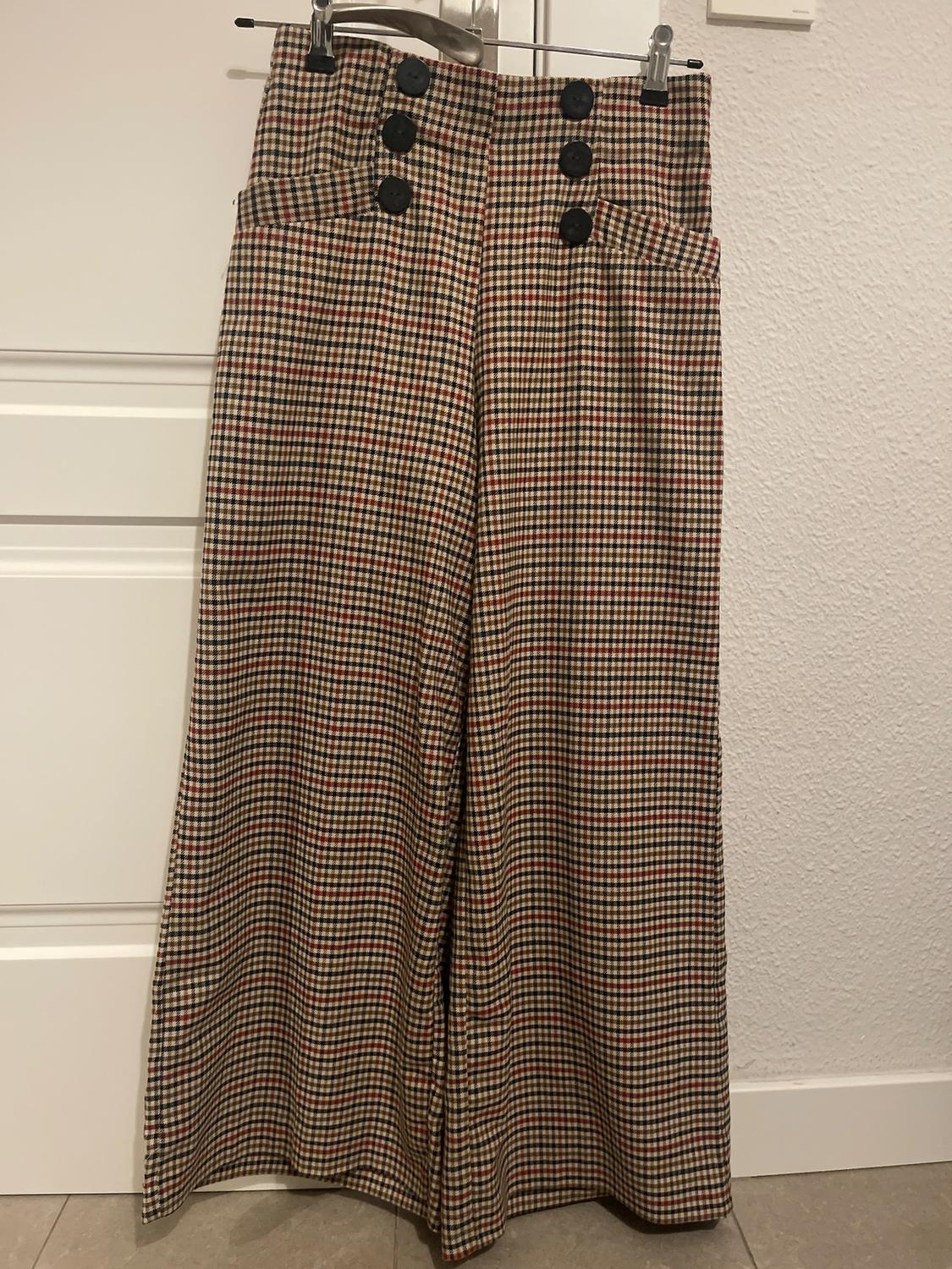Checked trousers with black buttons at the waist, hanging from a hanger over a white door.