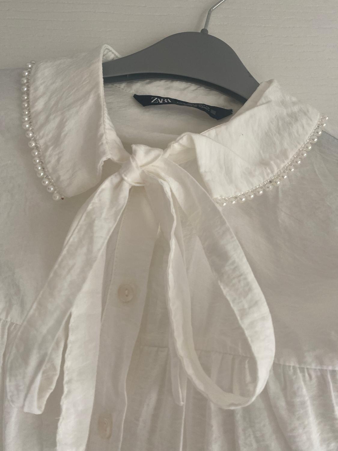 White blouse with a pearl-embellished collar and bow, hanging on a grey hanger. Visible label with text "ZARA".
