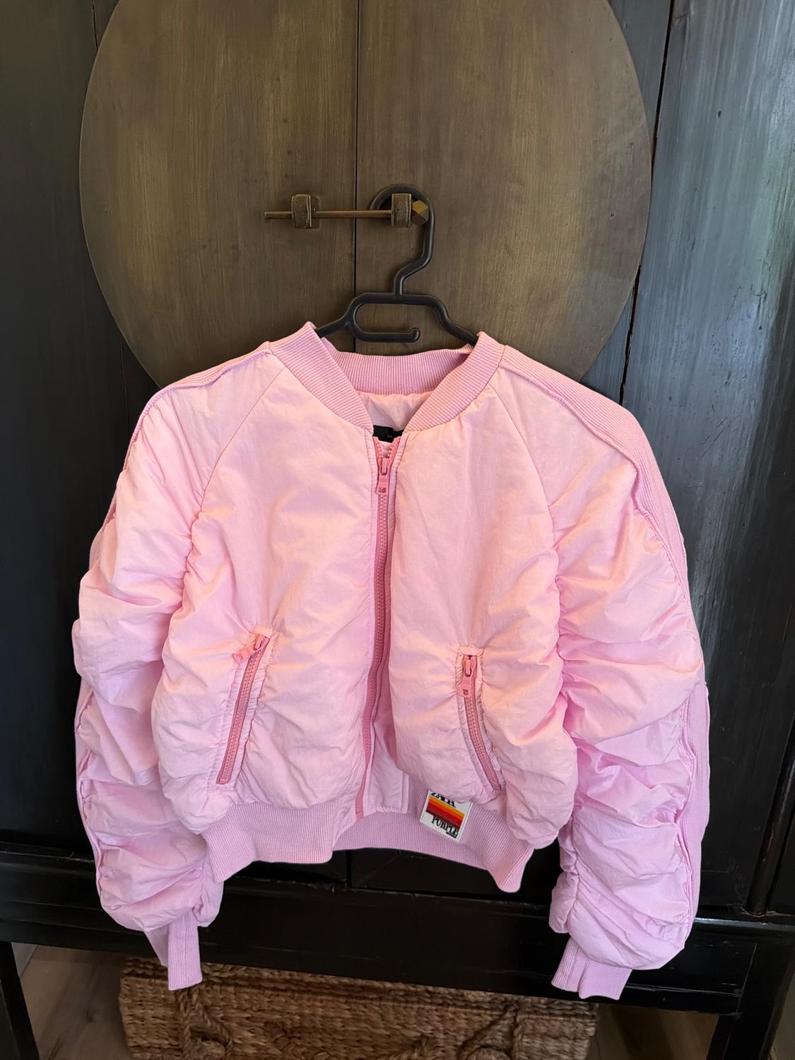 Pink bomber jacket with quilted texture, hanging on a black hanger in front of a dark wooden background.