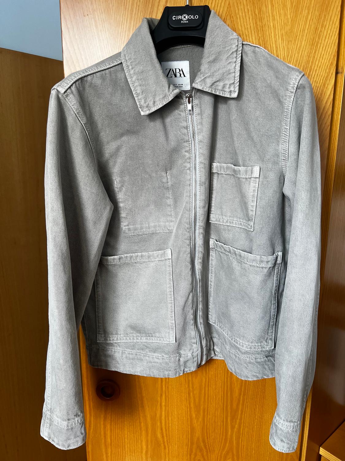 Grey fabric jacket with a zip and four front pockets, hanging on a black hanger with a "ZARA" tag.