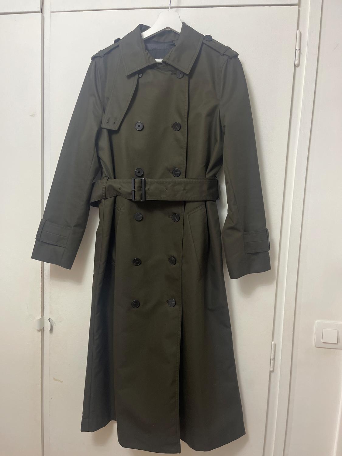 Olive green long coat with belt and black buttons, hanging on a white hanger in front of a white door.