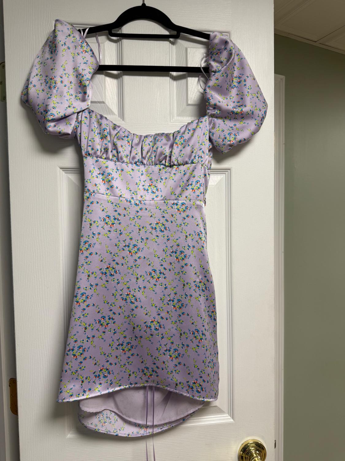 Lilac dress with a blue and green floral print, puff sleeves, hanging on a black hanger over a white door.