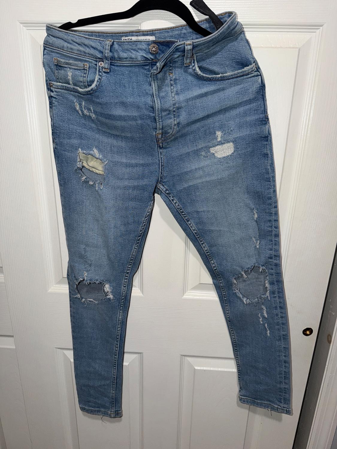 Worn blue jeans with rips at the knees and thighs, hanging on a black hanger over a white door.