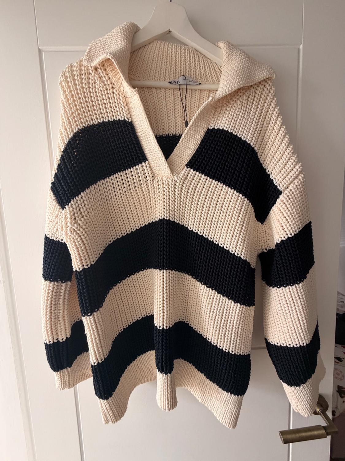 Chunky knitted jumper with horizontal black and beige stripes and a polo collar, hanging on a hanger on a white door.