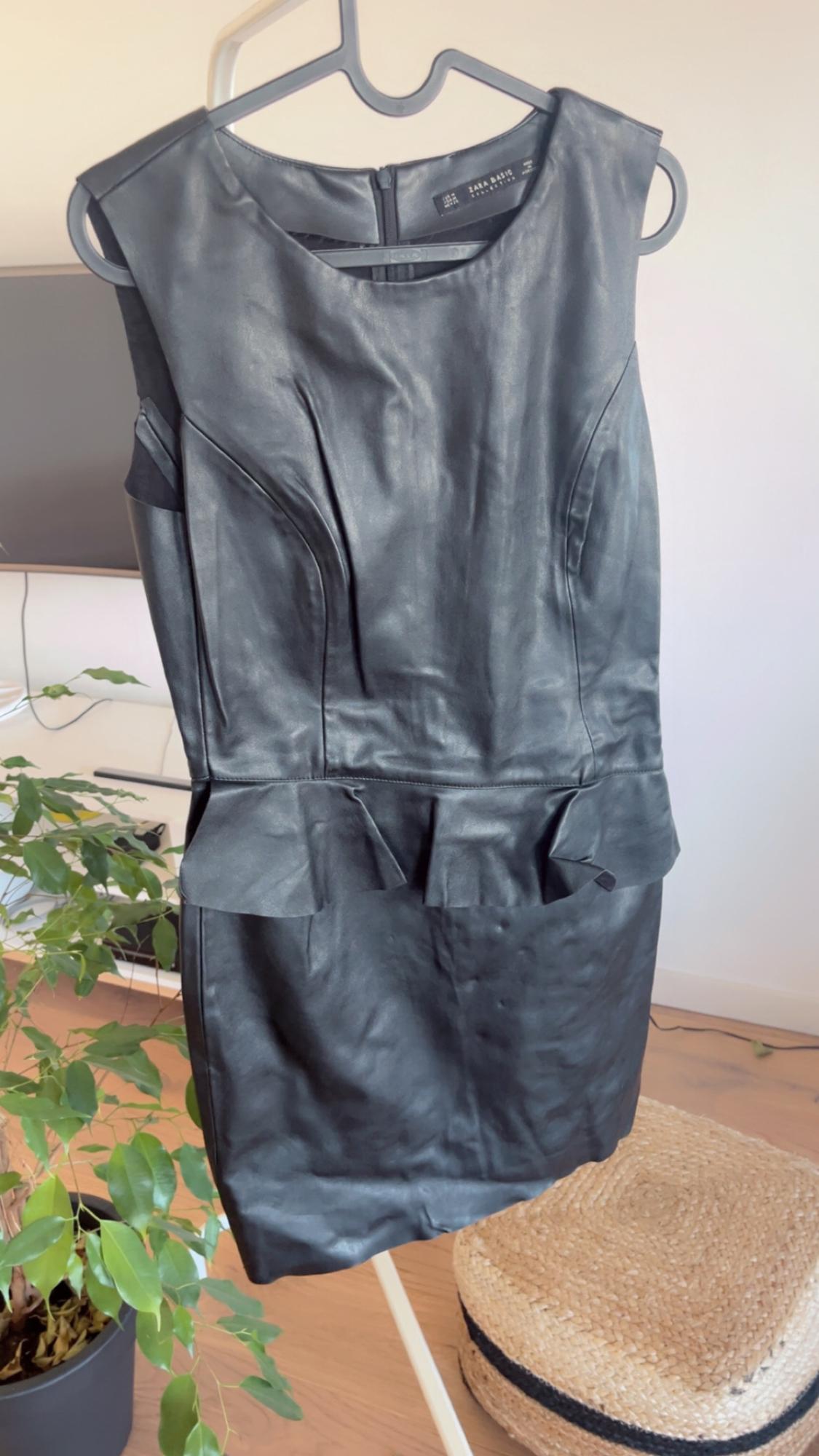 Sleeveless black dress hanging on a hanger, with frill details at the waist. Background with plant and wicker stool.