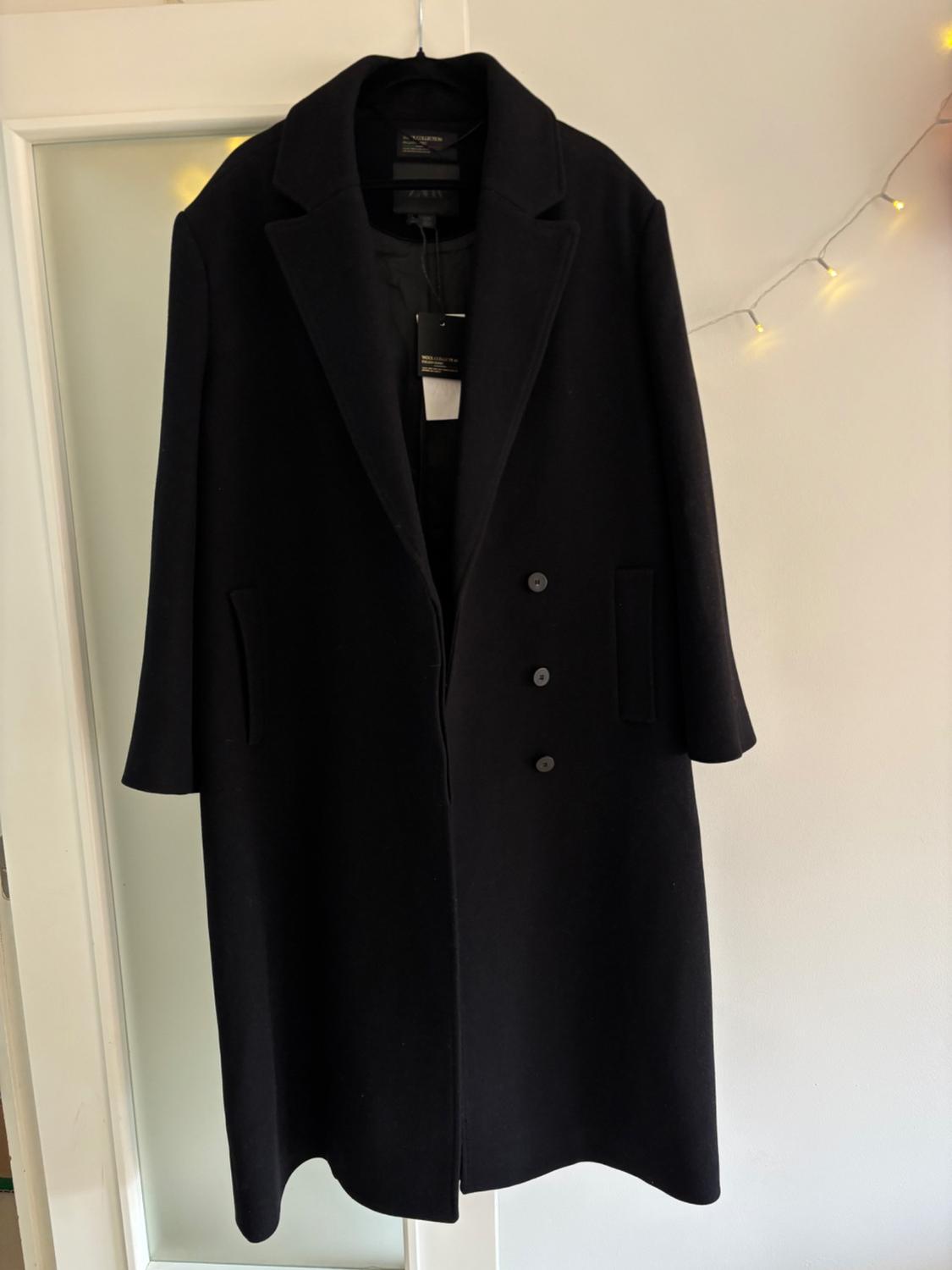 Black wool coat hanging on a white door, with decorative lights in the background. Label visible inside.