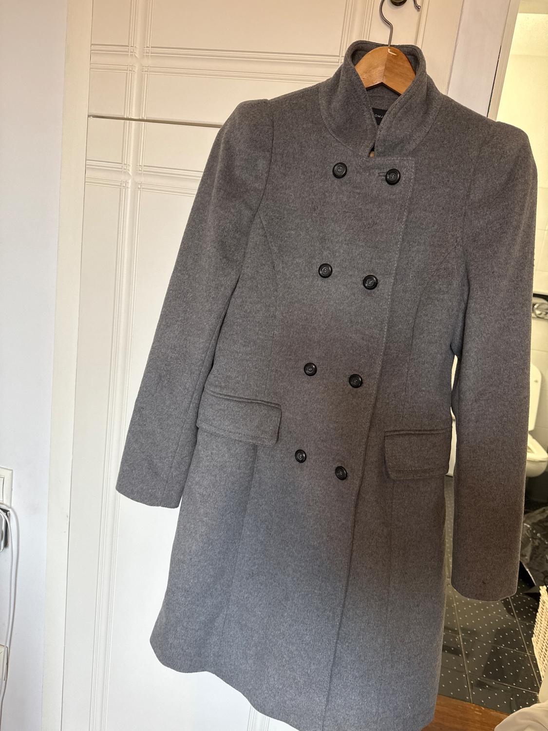 Grey wool coat hanging on a wooden hanger, with double-breasted fastening and pockets, against a white door background.