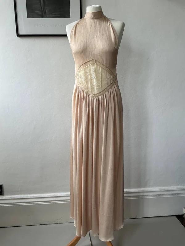 Mannequin in a long beige dress, with a halter neck, pleated skirt, and lace panel at the waist. White wall background with a framed picture.