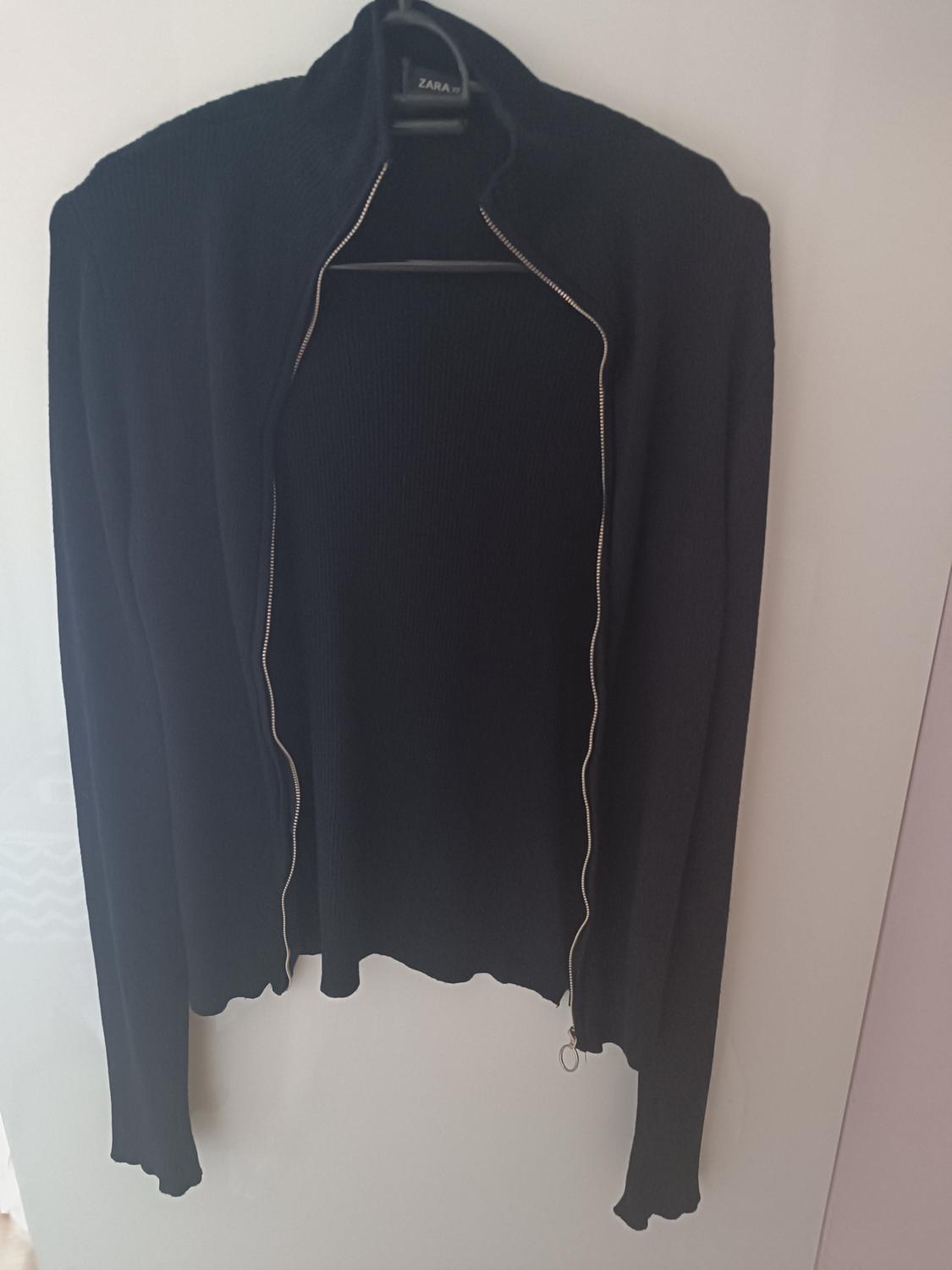Black knitted jacket with an open zip, hanging on a hanger with the "ZARA" brand visible on the collar.