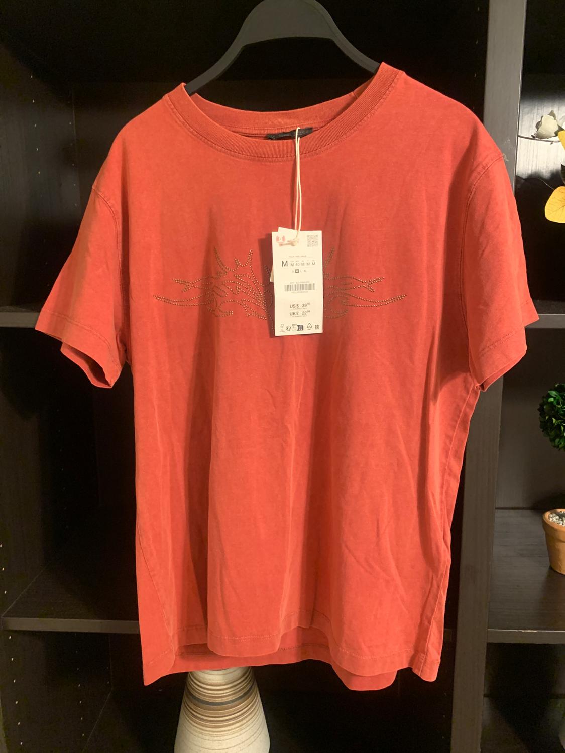 Red t-shirt hanging on a black hanger, with visible price tag. Dark shelf background with minimalist decor.