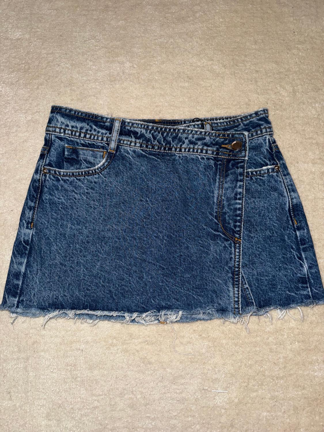Blue denim mini skirt with frayed hem on a beige background. Classic design with pockets and button fastening.