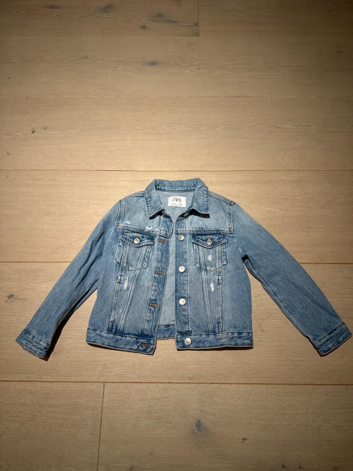 Light blue denim jacket with metal buttons and front pockets, on a light wood floor. Visible label on the collar.
