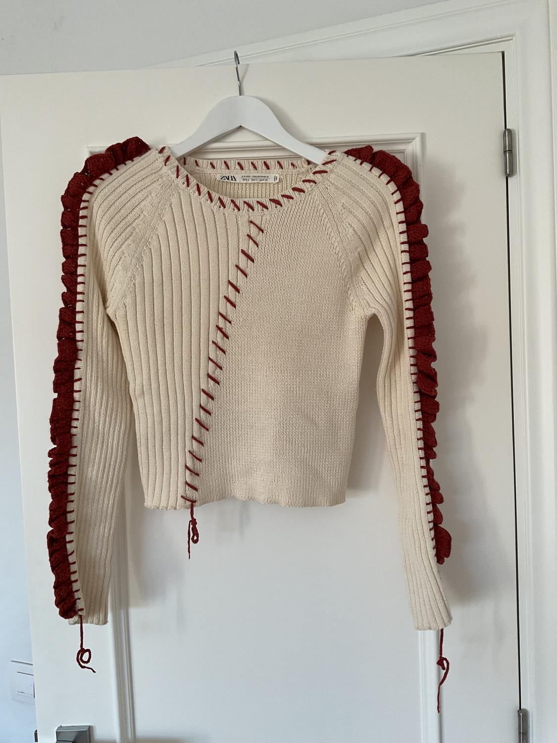 Beige knitted jumper with red details on the edges and seams, hanging on a white hanger over a white door.