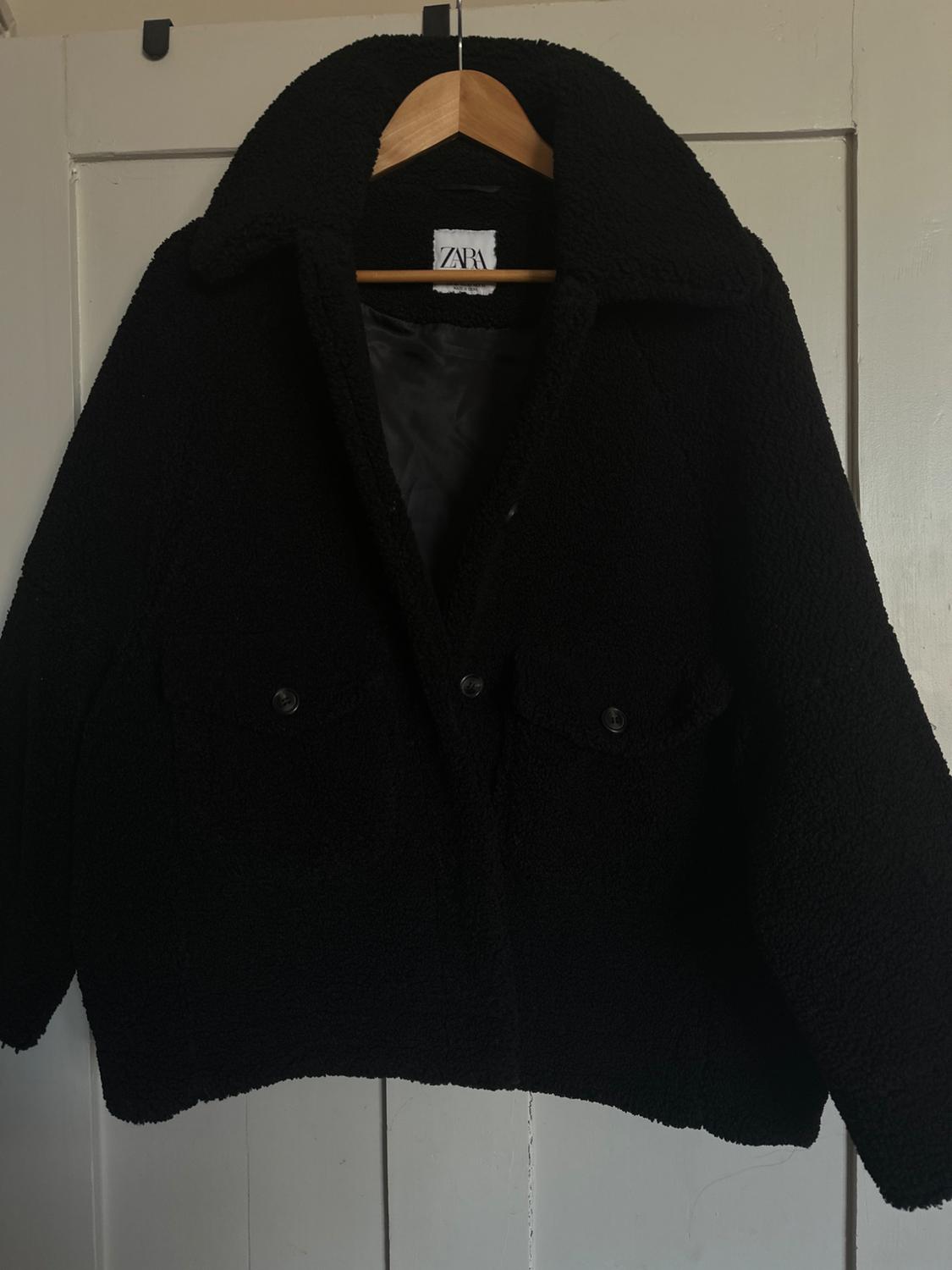 Black wool jacket hanging on a wooden hanger, with a visible label that says "ZARA", over a white door.