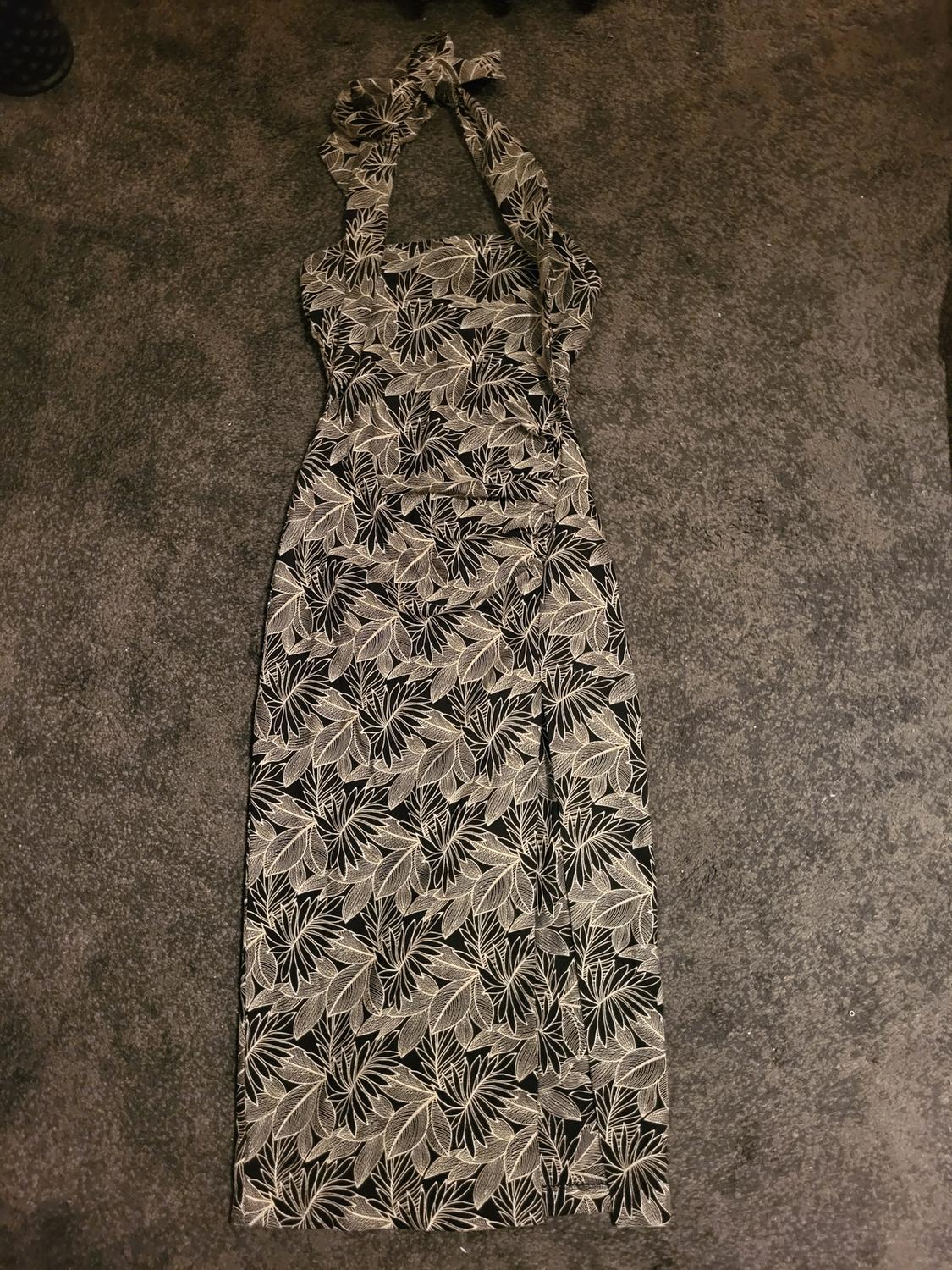 Long dress with a black and white leaf print on a dark background, with wide straps and a neck tie, on a grey carpet.