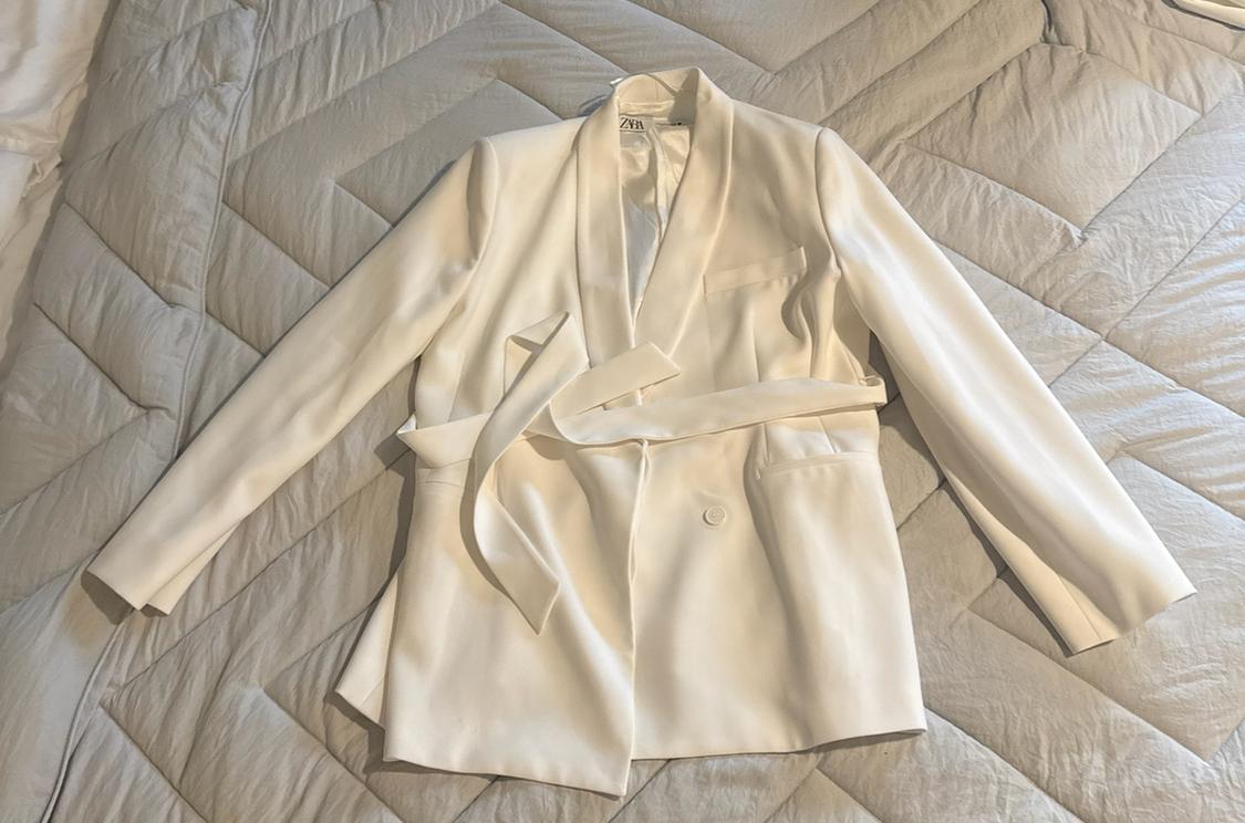 White jacket with a tied belt and ZARA label, laid out on a light grey bedspread. Relaxed style composition.