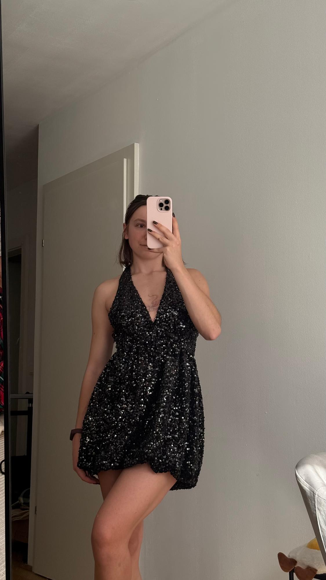 Woman in a shiny black dress takes a selfie in front of a mirror in a room with light walls and a white door.