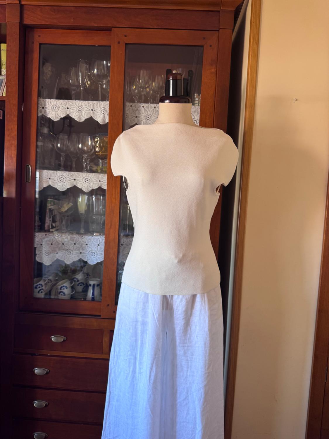 Mannequin wearing a white short-sleeved top and light-coloured flowing trousers, posing in front of a classic wooden display cabinet with glassware.