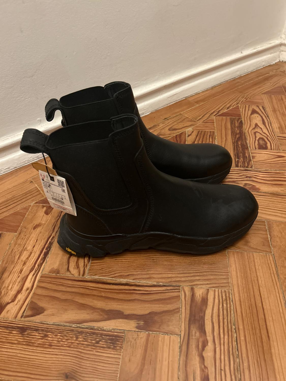 Black leather boots with thick soles on a wooden floor. Hanging tag visible on one of the boots.