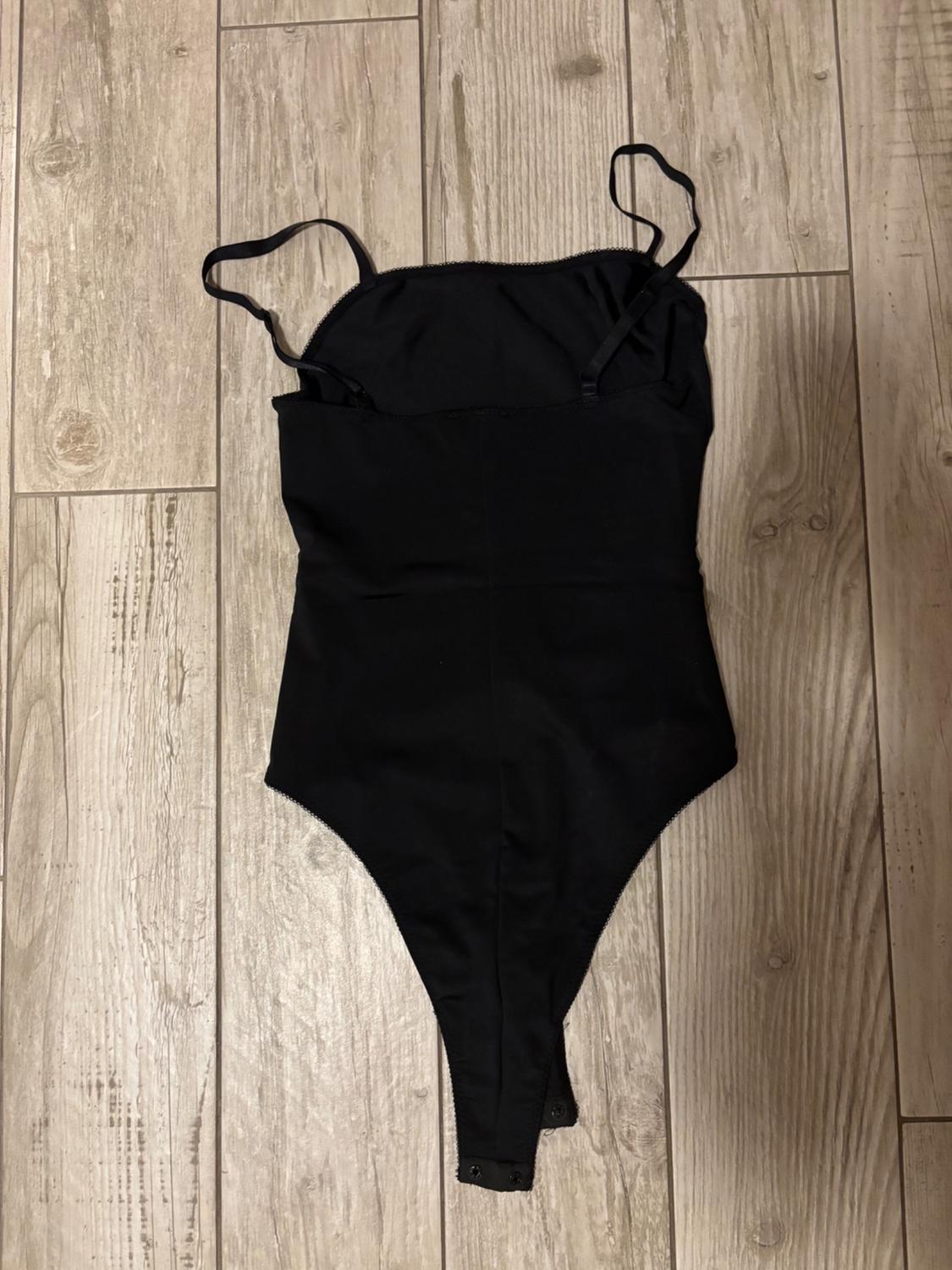 Black bodysuit with thin adjustable straps, viewed from behind and laid out on a light wooden floor.