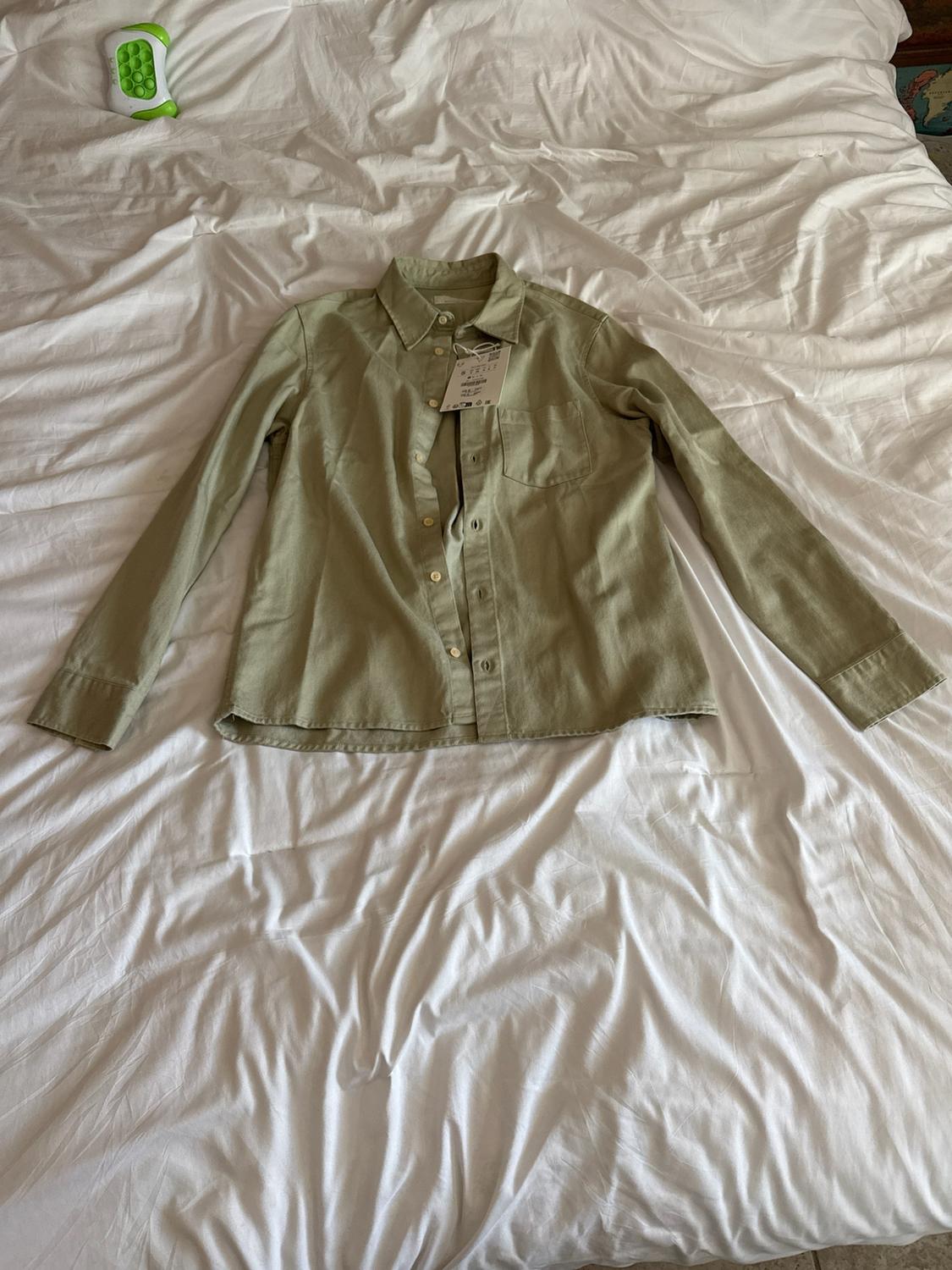 Light green long-sleeve shirt with front buttons and a pocket, laid out on wrinkled white sheets on a bed.
