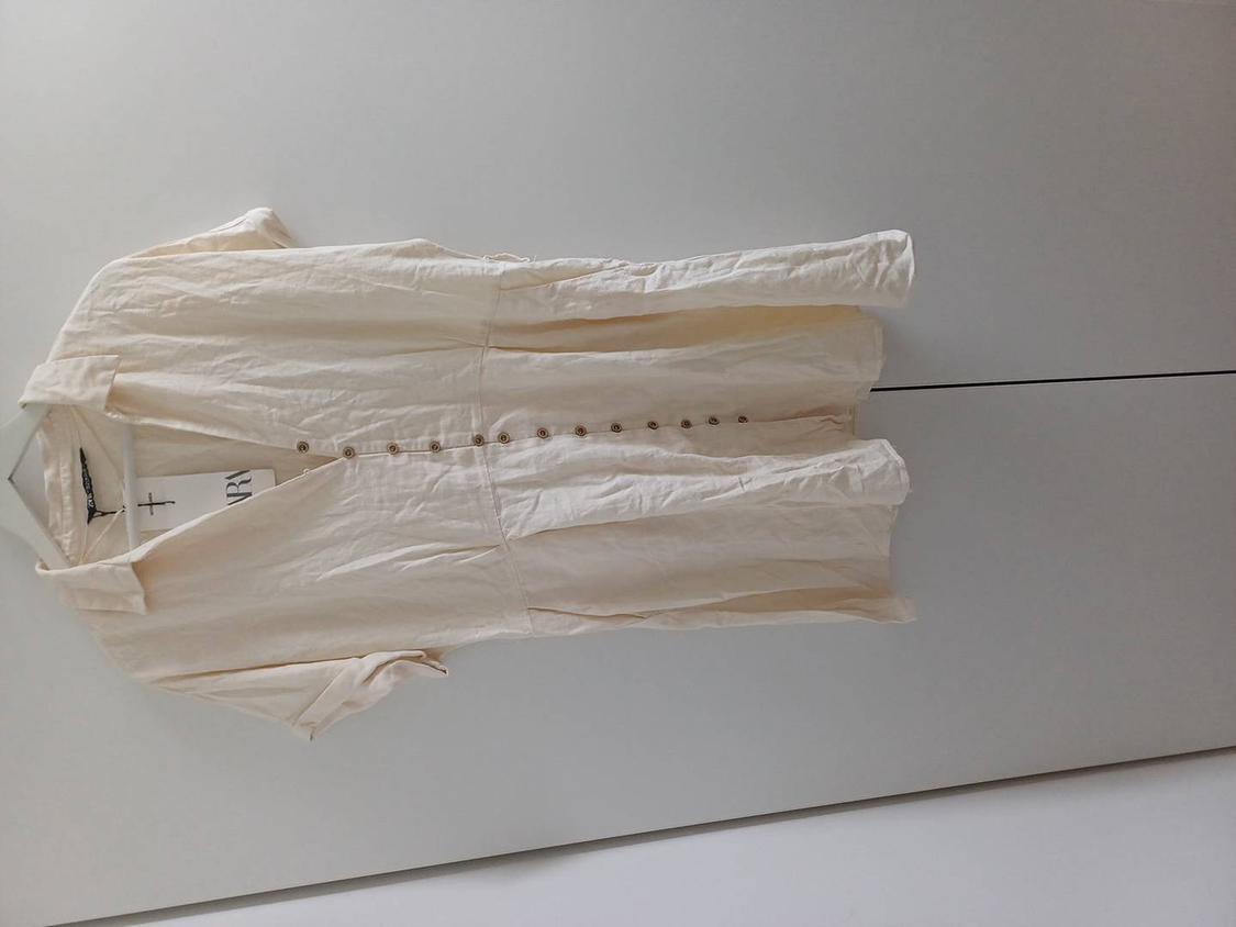 White linen jumpsuit with brown buttons hanging on a white hanger against a white background. Visible text: "ARY".