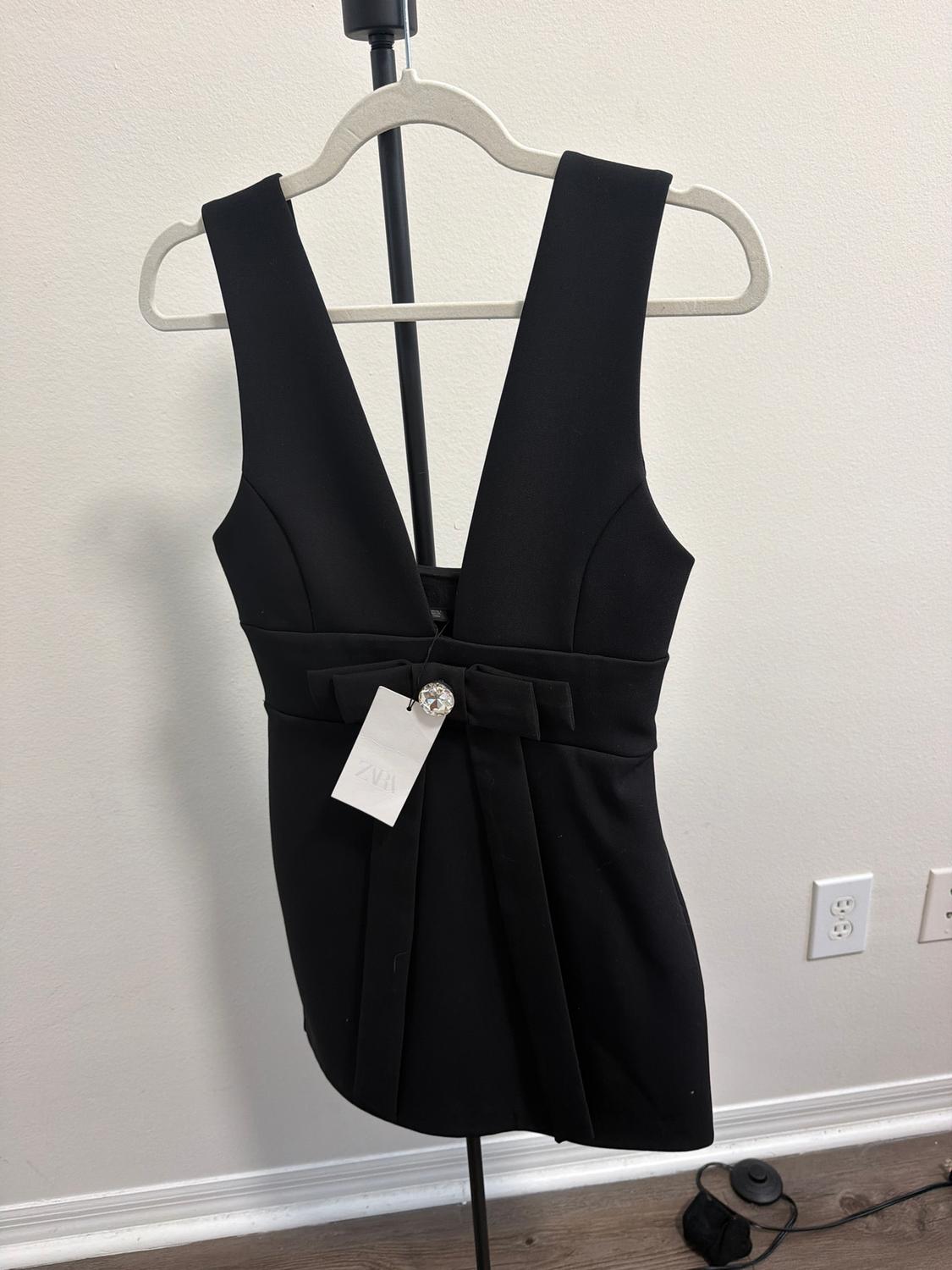 Sleeveless black dress on a hanger, with a bow at the waist and a hanging tag. White background and wooden floor.
