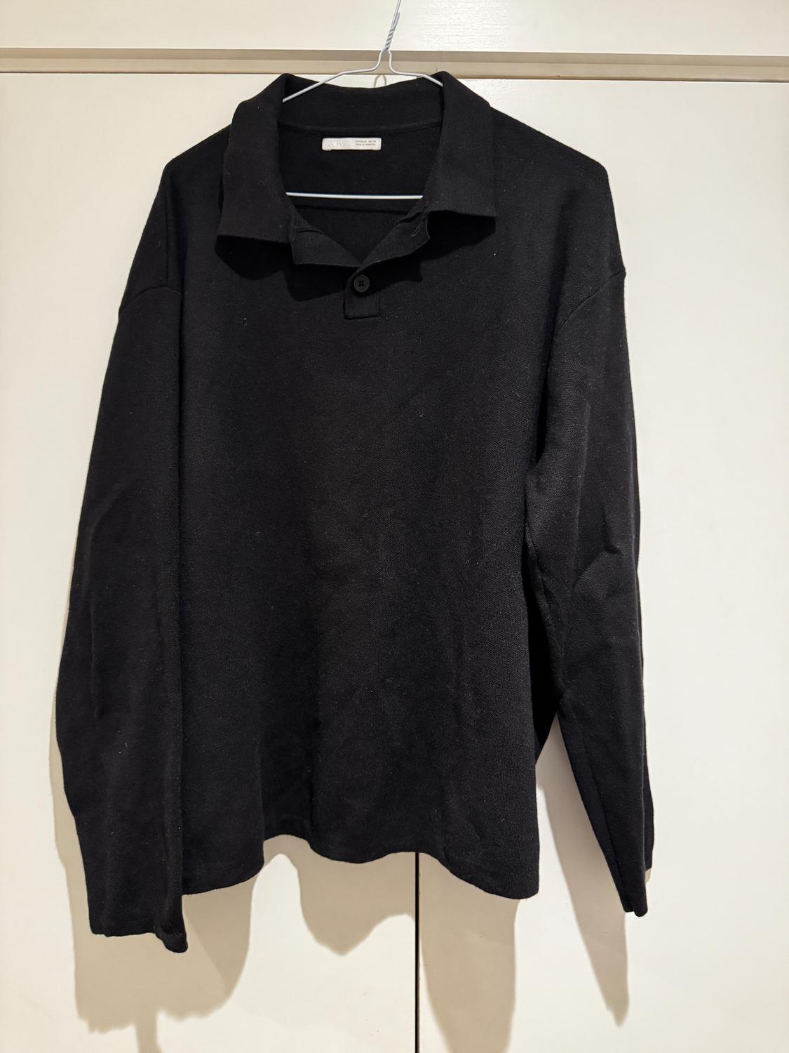 Long-sleeved black shirt hanging on a white hanger against a light background. It has a button on the collar.