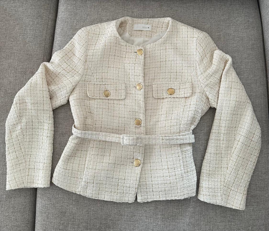 Cream textured fabric jacket with gold buttons and a belt, on a grey sofa.