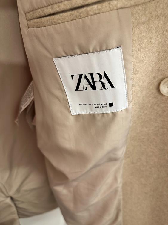 Beige garment tag with the "ZARA" logo. Text: "EUR L-XL USA L-XL MEX 42-44 MADE IN CHINA". Soft fabric and visible stitching.