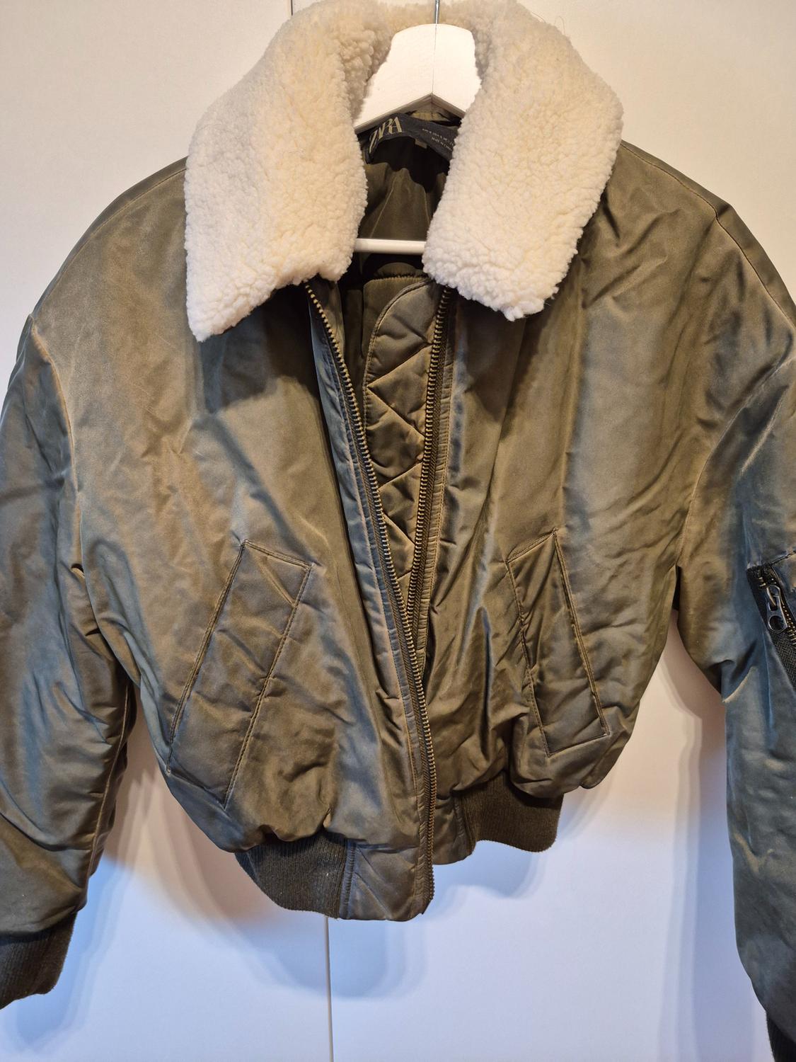 Olive green bomber jacket hanging on a white hanger. Its white collar, front zip, pockets and the ZARA label stand out.