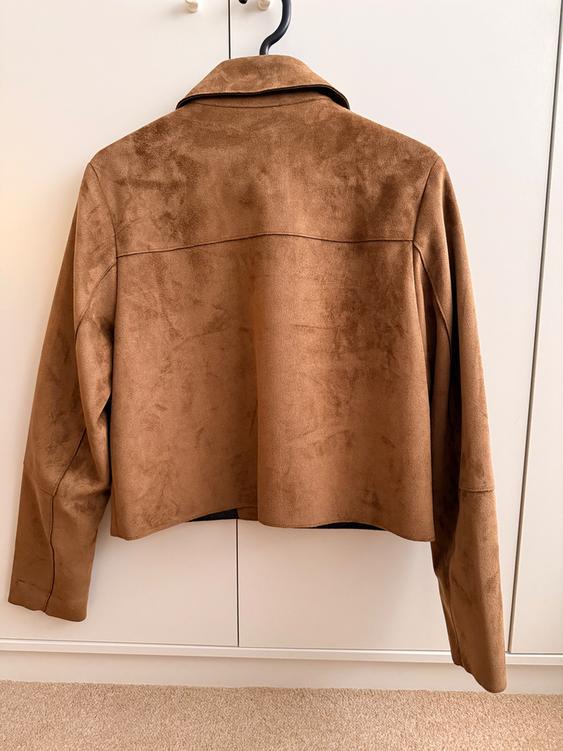 Brown suede jacket hanging on a black hanger in front of a white wardrobe. The jacket has a simple and elegant design.