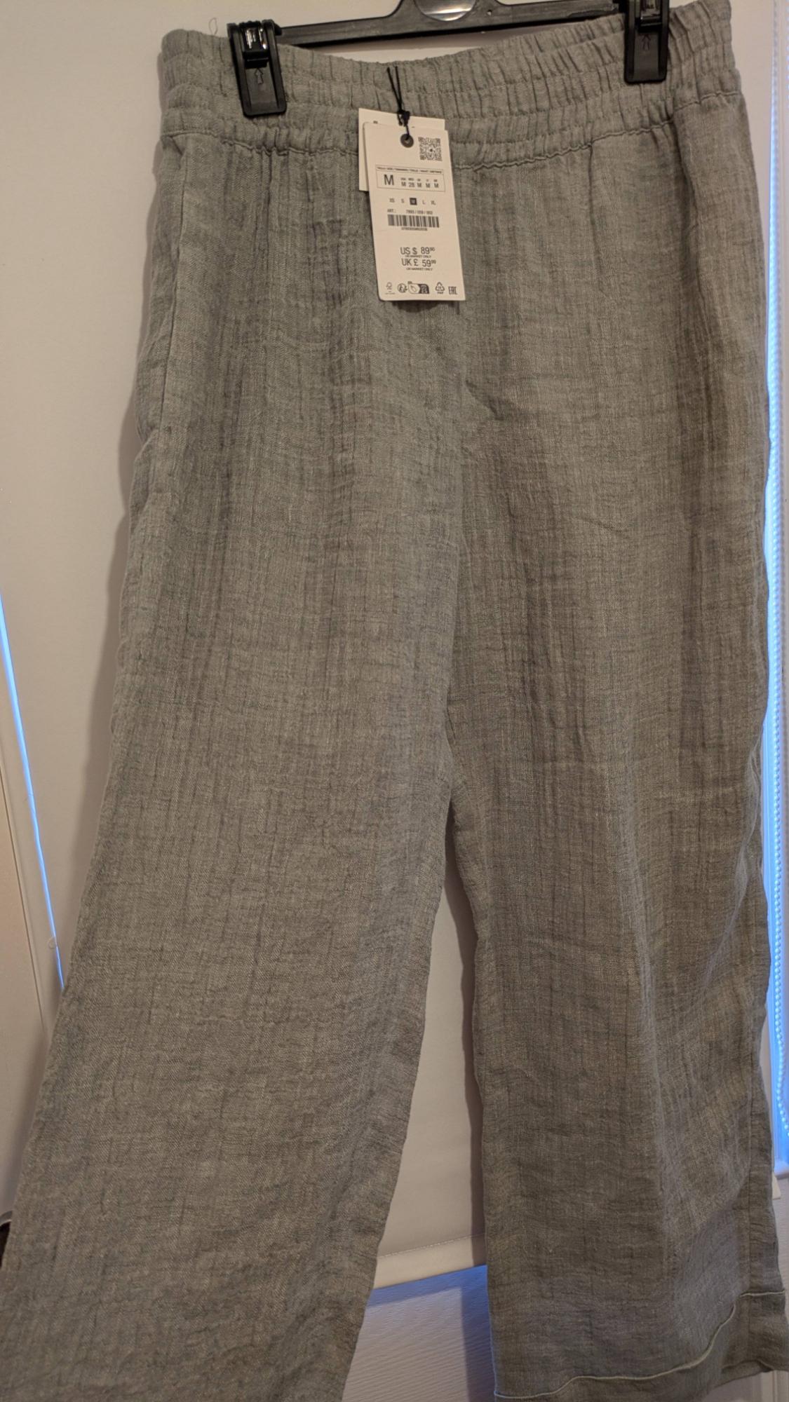 Light grey fabric trousers hanging on a hanger, with a visible price tag indicating size M and price US$ 8.99, UK £ 5.99.