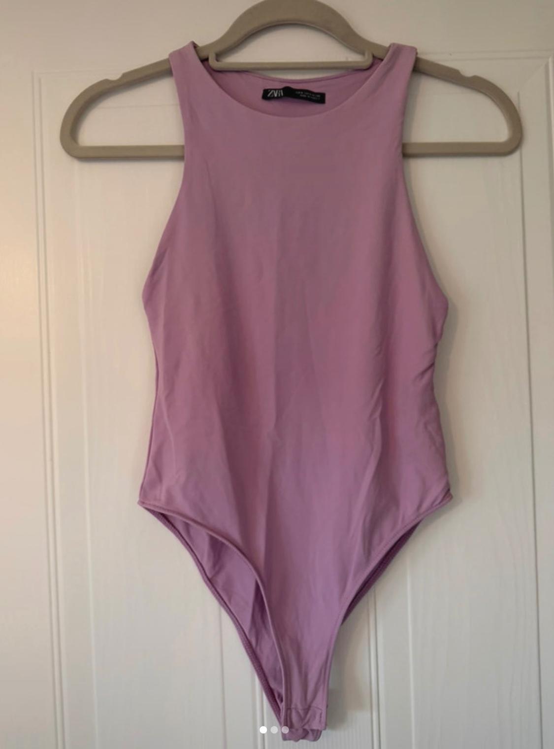 Lilac sleeveless bodysuit hanging on a hanger against a white door. The label with the text ZARA stands out on the neck.