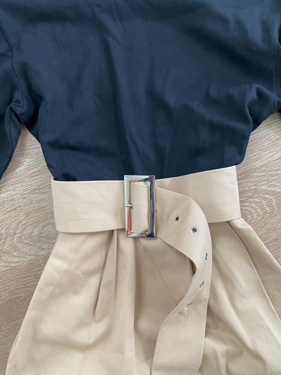 Black shirt paired with a high-waisted beige skirt, cinched with a wide belt with a metallic buckle against a light wood background.