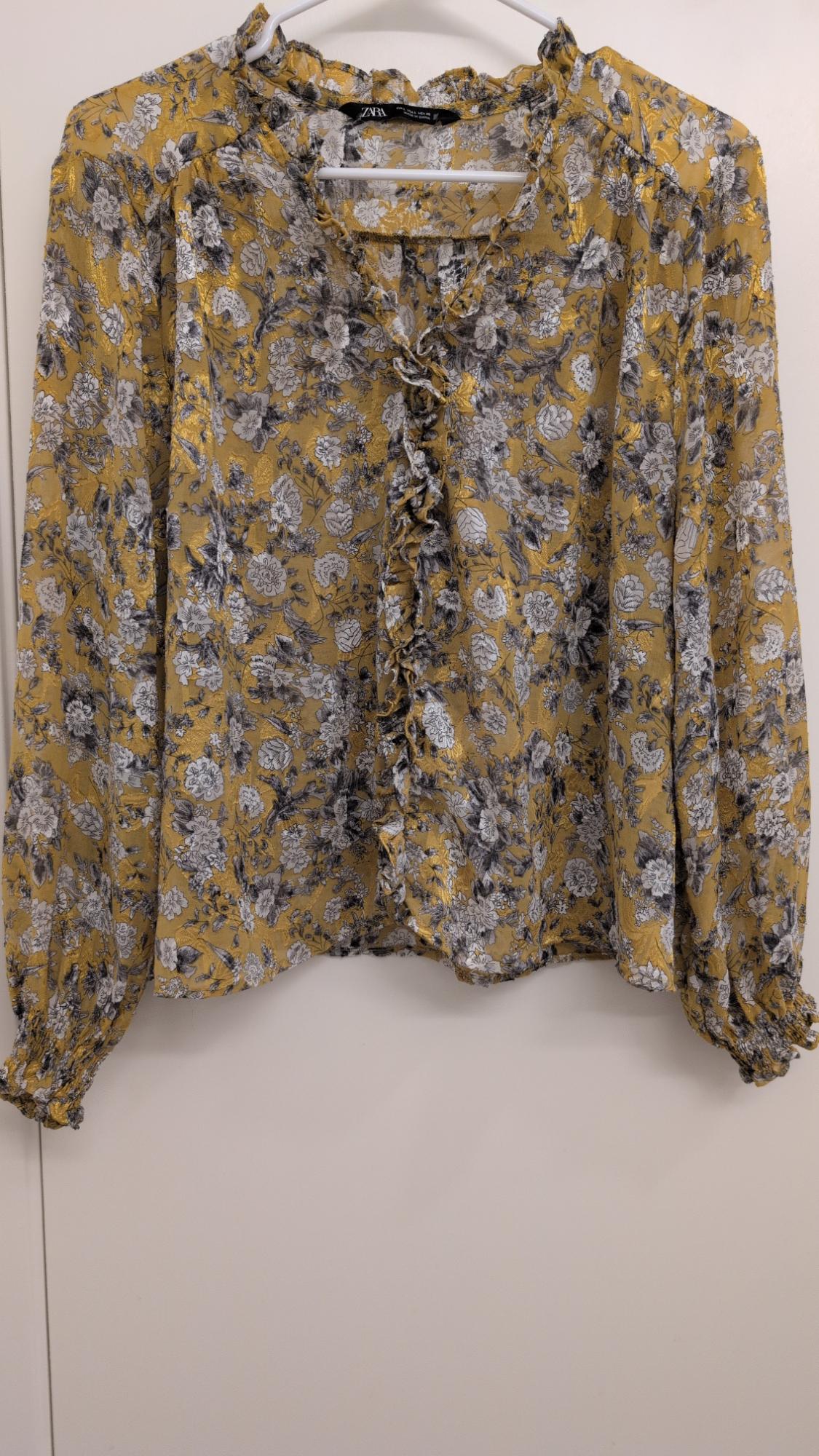 Yellow blouse with a black and white floral print, hanging on a white hanger. Ruffle detail on the front.