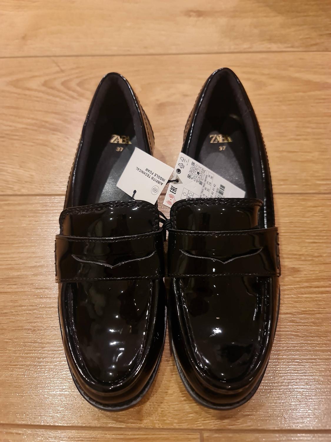 Black patent leather loafers on a wooden floor. Visible text: ZARA 37, AIRFIT® TECHNICAL INSOLE FOAM, EAC.