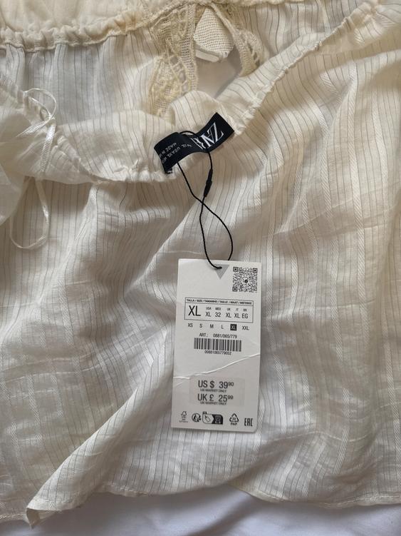 White fabric with fine stripes and a hanging tag showing size XL and prices: US $39.90, UK £25.99. Black tag with text.