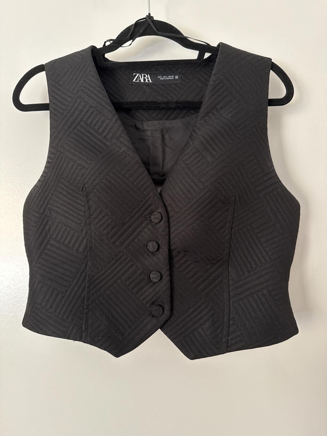 Black waistcoat with a geometric texture and front buttons, hanging on a black hanger against a white background. Interior label with the text ZARA.
