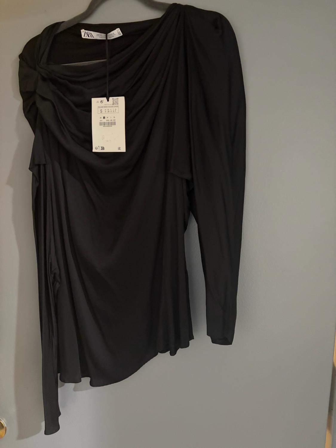 Asymmetrical, draped black long-sleeve top hanging on a hanger against a gray background. Visible tags with the text ZARA and S.