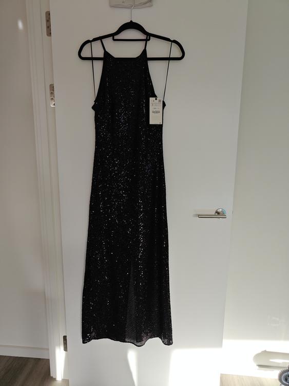 Black sequined dress hanging on a black hanger over a white door. Label visible on the right side of the dress.