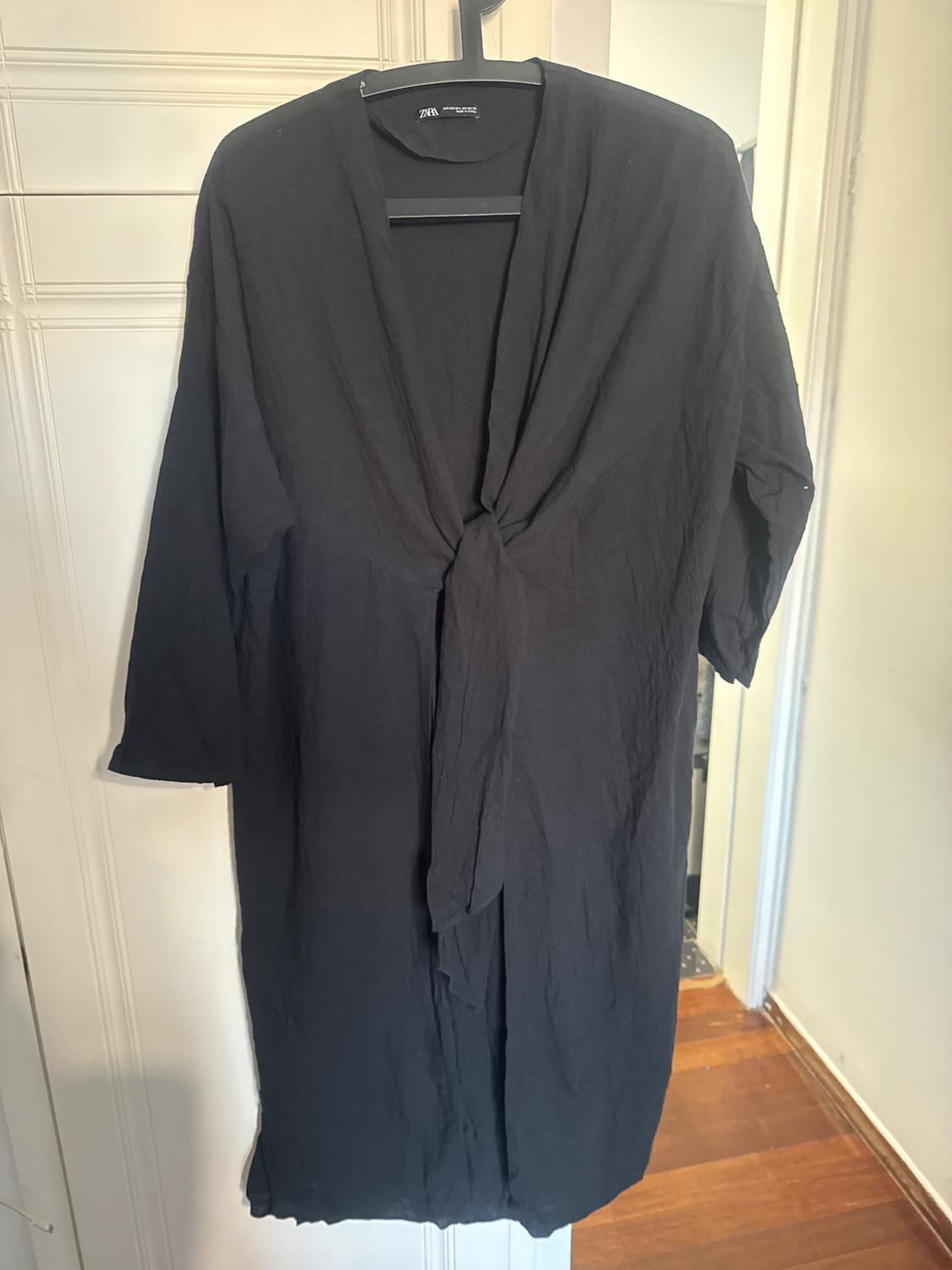 Black, long-sleeved dress hanging on a hanger, with a knot at the front. White wall background and wooden floor.