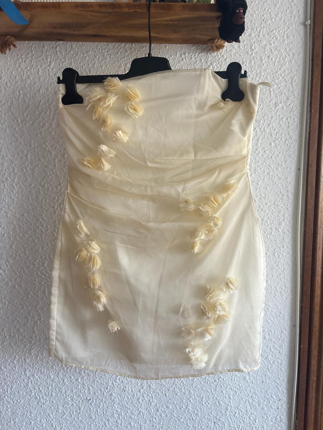 Cream bandeau mini dress with draping and embossed flowers, hanging on a black hanger against a white wall.