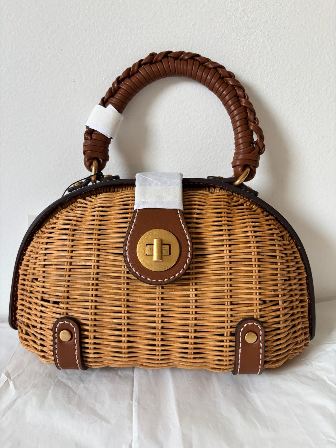 Wicker handbag with a braided brown handle, gold-tone metal closure, and protective paper details, resting on a white background.