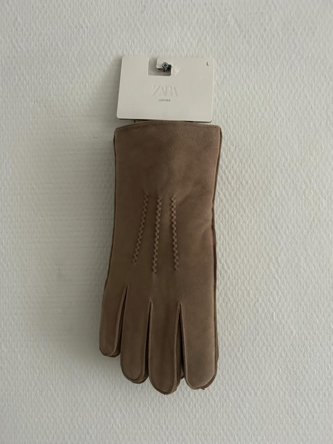 Brown glove hanging on a white wall with a label that reads "ZARA LEATHER L". Textured background.