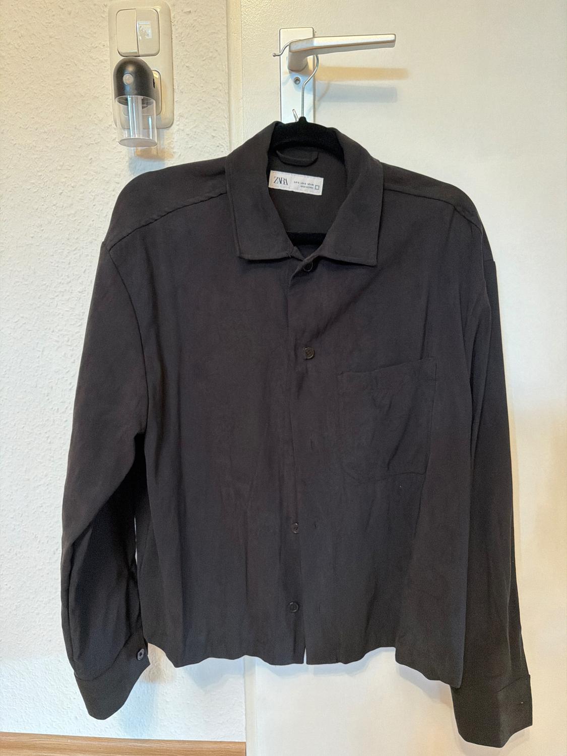 Black long-sleeved shirt hanging on a hanger over a white door. Visible label with text "ZARA". White wall background.